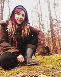 Frau mit gefärbtem Haar in brauner Jacke und Mütze hockend im Wald