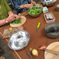 Hände zerkleinern frische Kräuter an Holzbrett; Schüsseln, Messer und Zutaten auf Tisch.