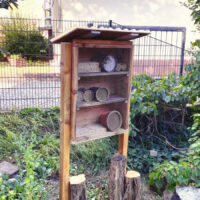 Ein Holzkasten mit drei Regalfächern, die verschiedene durchlöcherte Holzstücke, dünne Röhrenbündel und Töpfe mit Lehm enthalten